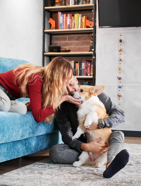 Young pair caressing their pet at home in modern living room. Husband sitting on rug and hugging dear dog, wife sitting on blue sofa and leaning to him petting under chin.