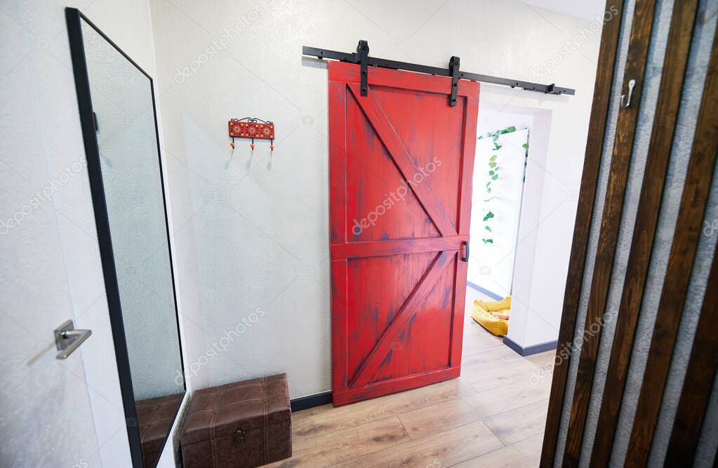 View of red door in corridor with big mirror on wall and ottoman. Interior of hallway of appartment in loft style with wooden flooring. Concept of house decoration.
