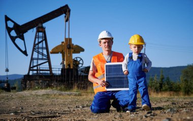 Yelek ve kask giyen mühendis güneş panelini çocuğun yanında tutuyor. Tecrübeli tamirci ve onun küçük takipçisi petrol kulesine karşı. Güneş enerjisi petrol endüstrisine ekolojik alternatiftir.