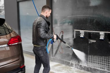 Araba yıkama istasyonunda araba yıkayan genç bir adam. Yüksek basınçlı su kullanarak araba temizleyen yakışıklı bir sürücü..
