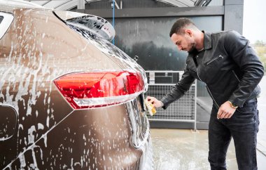 Dışarıdaki oto yıkama servisinde araba yıkayan sıradan giysili hoş bir adamın yan görüntüsü. Adam sabunlu süngeri aldı ve arabanın bagajına doğru eğildi. Plakasını süngerle siliyordu..