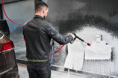 Arabaları yıkamak için yüksek basınçlı su tabancası tutan bir adam. Lastik araba paspaslarına köpük deterjan koyan adam araba yıkama duvarındaki metal ızgaraya sabitlenmiş..