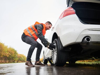 Sokakta araba tamir ederken yol kenarı yardım görevlisi lastiği söküyor. Yelekli erkek tamirci yolda lastik değiştiriyor. Acil yol hizmeti kavramı.