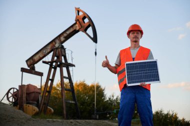 Elinde güneş paneli olan ciddi bir mühendisin portresi mavi gökyüzünün altındaki petrol pompa kulesinin arka planında sınıfını gösteriyor. Çevre dostu fotovoltaik enerji kavramı.
