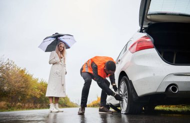 Erkek oto tamircisi, şık bir kadın şemsiyesini tutarken direksiyonda fındık söküyor. Yelekli genç adam yolda kadın otomobili tamir ediyor. Acil yol hizmeti kavramı.