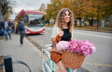 Sepetinde çiçeklerle bisiklete binmiş güzel genç bir kadın. Onay jesti yapıyor. Şehrin göbeğinde bisiklet sürerken gülümseyen neşeli kadın..