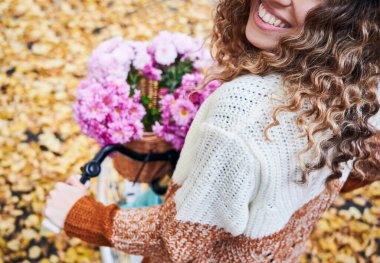 Sepetinde bir buket çiçekle bisiklete binen kıvırcık saçlı neşeli genç bayanı yakından takip edin. Güzel gülüşü olan, elinde pembe çiçeklerle bisiklet gidonu tutan bir kadın. Gülümseyen kadına odaklan..