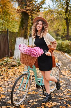 Önde, bej şapkalı, mutlu gülümseyen bir kadın, şirin mavi bisikletli, kahverengi sepetli ve pembe güzel çiçekli. Parkta şık bir bisikletle yürüme kavramı.