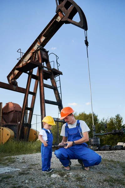 Engineer father introducing his son to work tool. Adult man and preschool child in workwears and helmets together looking to adjustable wrench on background of pump for oil production.