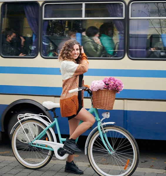 곱슬곱슬 머리의 여자가 자전거를 파란색 흰색으로 뒷자리에 꽃다발처럼 분홍색 들어온다 — 스톡 사진