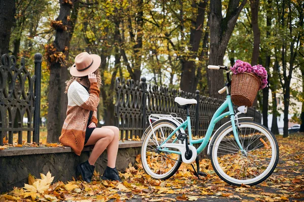 城市秋季公园里 身穿米黄色帽子 穿着时髦衣服的年轻女子坐在蓝色的现代自行车旁边 提着篮子鲜花的侧视图 公园内新鲜空气步行的概念 — 图库照片