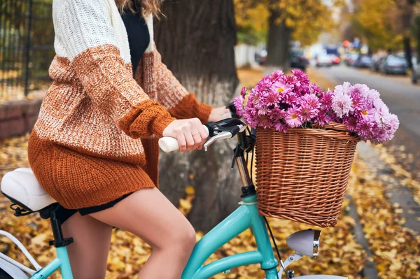 Close up of female bicyclist in knitted cardigan riding bike with flowers on autumn street. Young woman on bike with beautiful bouquet in front basket.