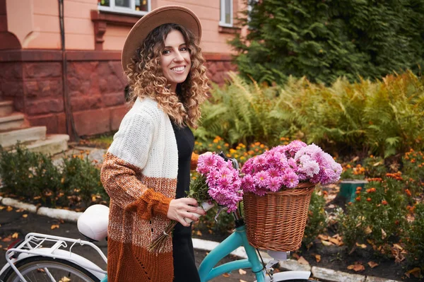 バスケットに花と自転車を保持しながら カメラと笑顔を見て巻き毛のきれいな女性 陽気若いです女性身に着けていますエレガントな帽子とカーディガンながら時間屋外 — ストック写真