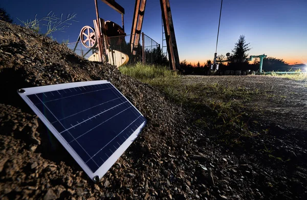 Close up view of one small solar panel, which located on stone mound. The lower part of oil well on the background. Concept of solar energy as alternative to oil production.