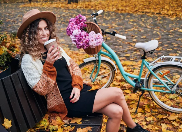 곱슬머리의 여자가 모자를 자전거 의나무 벤치에 공원에서 맛있는 뜨거운 커피나 — 스톡 사진