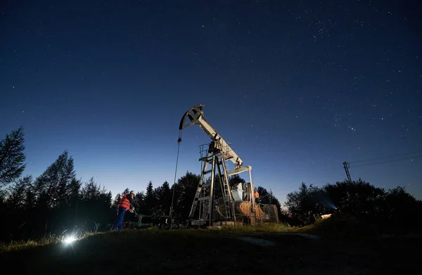 Night work of oil rig. Engineer correcting serviceability of the pipeline near rod pump. Silhouettes of trees and beautiful starry sky among petroleum derrick.