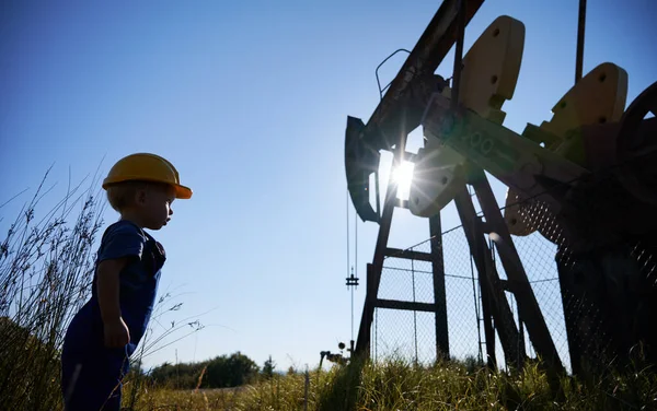 Side low view of child boy who standing opposite metal fence and examining oil pump structure in summer sunny day. On the background cloudless blue sky.