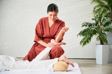 Masseuse making thai yoga massage. Woman therapist doing traditional massage treatment for female patient using elbow. Client lying on mat, meditating, enjoying hand massage in calm atmosphere.