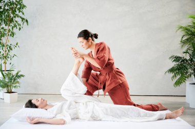 Masör Tayland yogası masajı yapıyor. Geleneksel masaj tedavisi uygulayan kadın terapist, kadın hastaların vücudunu geriyor. Müşteri paspasın üzerinde uzanıyor, sakin atmosferde el masajının keyfini çıkarıyor..