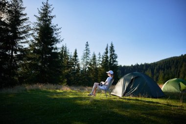 Kadın kampçı, fotoğraf makinesiyle çadırın yanında dinleniyor. Etrafı hep yeşil ağaçlarla çevrili ve yumuşak akşam ışığında yıkanan dişi turist, o anın huzurunu yakalıyor..