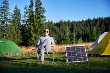 Çevre dostu kamplarda fotovoltaik güneş paneli kullanarak taşınabilir elektrik santralini şarj eden bir adam. Erkek turist çadırların yanındaki sandalyede rahatça oturur. Arka planda uzun çam ağaçları ve açık mavi gökyüzü.