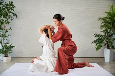 Masör Tayland yogası masajı yapıyor. Geleneksel masaj tedavisi uygulayan kadın terapist, kadın hastaların vücudunu geriyor. Müşteri paspasın üzerinde oturuyor, sakin bir atmosferde el masajının keyfini çıkarıyor..
