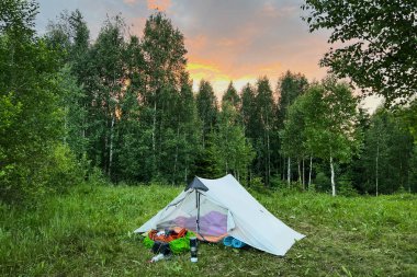 Beyaz turist çadırı, çimenli bir alanda sık ağaçların arkasında kamp malzemesi. Güneş batarken gökyüzü turuncu ve pembe parlak renklerle parlıyor. Huzurlu kamp alanının üzerine huzurlu bir ortam yaratıyor..