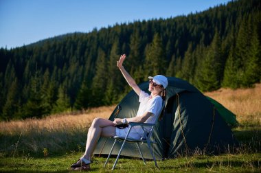 Kadın kampçı, fotoğraf makinesiyle çadırın yanında dinleniyor. Etrafı hep yeşil ağaçlarla çevrili ve yumuşak akşam ışığında yıkanan dişi turist, o anın huzurunu yakalıyor..