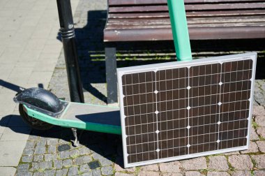 Nane yeşili elektrikli scooter, güneş panelinin yaslandığı kaldırım taşı yolunda duruyor. Güneş ışığını yakalamak için stratejik olarak yerleştirilen güneş paneli, scooter yüklemek için sürdürülebilir metodu vurguluyor.