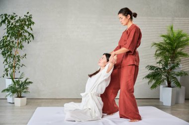 Masör Tayland yogası masajı yapıyor. Geleneksel masaj tedavisi uygulayan kadın terapist, kadın hastaların vücudunu geriyor. Müşteri paspasın üzerinde oturuyor, sakin bir atmosferde el masajının keyfini çıkarıyor..