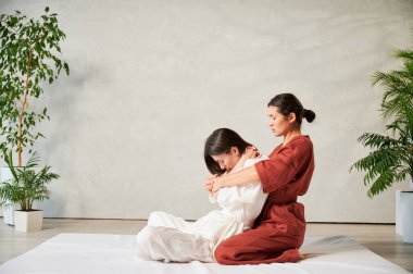 Masör Tayland yogası masajı yapıyor. Geleneksel masaj tedavisi uygulayan kadın terapist, kadın hastaların vücudunu geriyor. Müşteri paspasın üzerinde oturuyor, sakin bir atmosferde el masajının keyfini çıkarıyor..