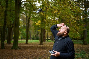 Sakallı bir adam verimli bir parkta sonbahar yapraklarıyla dolu bir drone kullanıyor. Açık hava deneyiminin tadını çıkarırken odaklanmış ve heyecanlı görünüyor..
