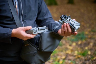 Eller dikkatlice katlanmış bir dronu ve kumandayı tutuyor, sakin bir ormanda düşen yapraklarla çevrili. Güneş ağaçların arasından süzülüp sükunet yaratır..