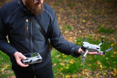 Bir adam bir elinde insansız hava aracını, diğer elinde de kumandayı tutar. Düşen yapraklarla dolu canlı bir parkta havalanmaya hazırdır..