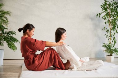 Masör Tayland yogası masajı yapıyor. Geleneksel masaj tedavisi uygulayan kadın terapist, kadın hastaların vücudunu geriyor. Müşteri paspasın üzerinde oturuyor, sakin bir atmosferde el masajının keyfini çıkarıyor..