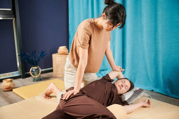 Masör Tayland yogası masajı yapıyor. Geleneksel masaj tedavisi uygulayan kadın terapist, kadın hastaların vücudunu geriyor. Müşteri paspasın üzerinde uzanıyor, sakin atmosferde el masajının keyfini çıkarıyor..