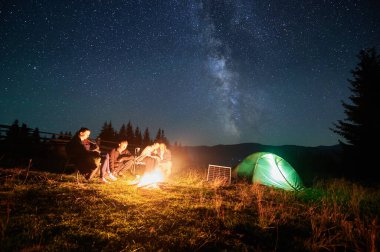 Yürüyüş yapan bir grup arkadaş kamp ateşi etrafında, yıldızlarla dolu gökyüzünün altında, aydınlık çadır ve güneş panelinin yanında dinleniyor. Samanyolu, dağ yabanda büyülü ve atmosfer yaratan parlak bir ışık saçar..
