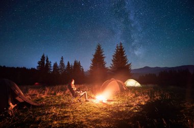 Yürüyüşçü kadın kamp ateşiyle gevşer. Ilık parıltısıyla aydınlanır. Yıldızlarla dolu gökyüzünün altında çadır kurar. Samanyolu başınızın üstüne uzanır ve dingin dağ düzenine sihirli bir dokunuş ekler..