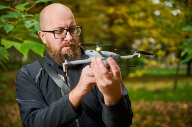 Sakallı ve gözlüklü bir adam renkli sonbahar yapraklarıyla dolu canlı bir parkta İHA 'yı yakından inceliyor. Sahne, uçuşa hazırlanırken onun odaklanmış yüz ifadesini yakalıyor..