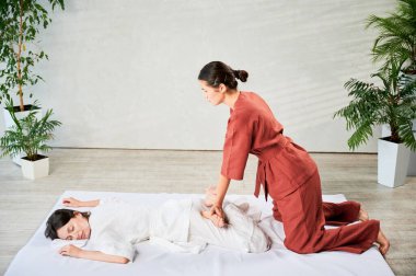 Masör Tayland yogası masajı yapıyor. Geleneksel masaj tedavisi uygulayan kadın terapist, kadın hastaların vücudunu geriyor. Müşteri paspasın üzerinde uzanıyor, sakin atmosferde el masajının keyfini çıkarıyor..