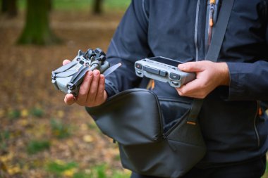 Bir insan bir elinde katlanmış bir dron, diğer elinde ise sakin bir ormanda doğayı keşfetmeye hazır bir kontrolör tutar..