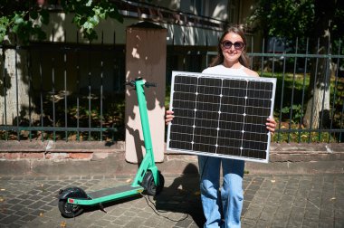 Mobilet yüklemek için taşınabilir güneş paneli kullanan kadın sürdürülebilir yenilenebilir enerji entegrasyonunu vurguluyor. Güneş paneline bağlı yeşil elektrikli scooter, çevre dostu ulaşımı vurguluyor.