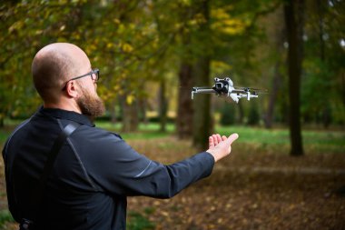 Sakallı bir adam renkli sonbahar ağaçlarıyla çevrili bir parkta küçük bir drone fırlatmaya hazırlanıyor. Bu yenilikçi teknolojiye odaklanırken toprağı kaplıyor..