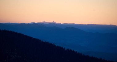 Gün batımında uzak dağların silueti, altın gökyüzü derin mavi gölgelere dönüşüyor. Sırtlar ve zirveler ufkun sıcak renklerine karşı çarpıcı bir tezat yaratır..