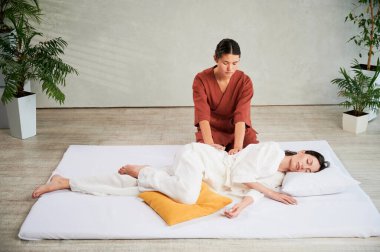 Masör Tayland yogası masajı yapıyor. Kadın terapist, kadın hasta için evde geleneksel masaj tedavisi uyguluyor. Müşteri paspasın üzerinde uzanıyor, meditasyon yapıyor, sakin bir atmosferde el masajının keyfini çıkarıyor..
