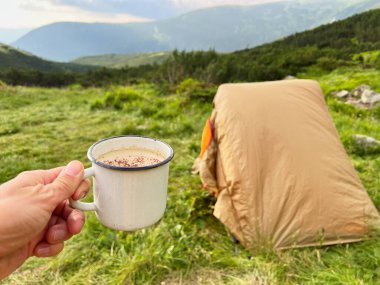 Beyaz diş minesi bardakları sıcak içecekle dolu, kakao tozlu, çimenli çadır üzerine kurulmuş bej bir çadırın önünde. Arkaplanda yuvarlanan yeşil tepeler ve parçalı bulutlu gökyüzü.
