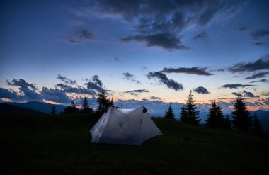 Alacakaranlıkta, dağların ve çam ağaçlarının karanlık siluetleriyle çevrili çimenli yamaçta turistik çadır sıcacık parlar. Gün batımının son ışığı kararırken gökyüzü koyu mavi ve dağınık bulutlarla kaplı..
