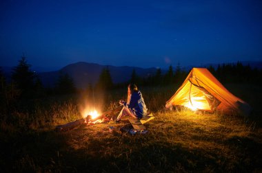 Kadın yürüyüşçü, sakin dağ manzarasıyla çevrili alacakaranlığın yumuşak parıltısıyla aydınlık çadırının yanında kamp ateşinin yanında oturur. Yukarıda açık bir gece gökyüzü sessiz vahşi doğaya huzur dolu bir ortam katıyor..