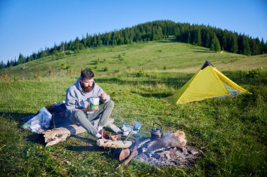 Sakallı adam yemek kesesini yiyor. Gri kapüşonlu ve yeşil pantolonlu turist kamp ateşinin yanında oturuyor. Yanında, kamp malzemeleri ve sarı çadır manzaralı açık hava ortamını ekleyin..