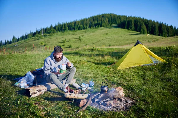 Sakallı adam yemek kesesini yiyor. Gri kapüşonlu ve yeşil pantolonlu turist kamp ateşinin yanında oturuyor. Yanında, kamp malzemeleri ve sarı çadır manzaralı açık hava ortamını ekleyin..